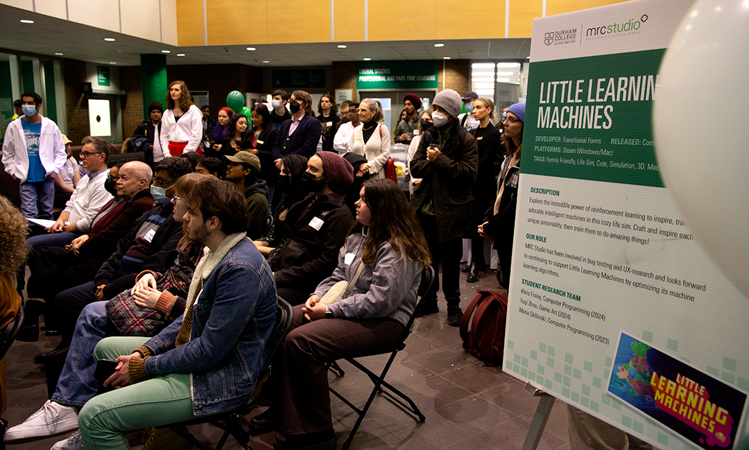 The crowd listens to the opening remarks at the MRC Arcade at Durham College. Little Learning Machines, shown on the sign, was one of five games created in the studio showcased at the event.