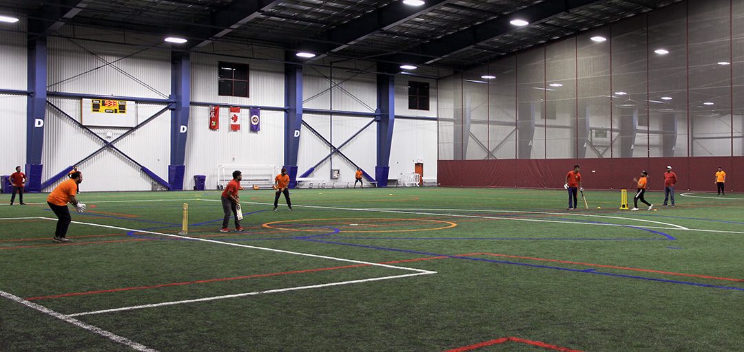 The Oshawa Cricket League match taking place between Team Red and Team Orange. The bowler on Team Orange winds up a throw towards the batsman on Team Red.