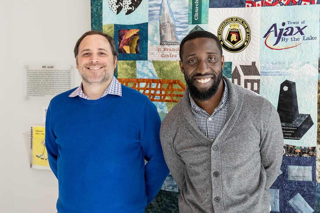 Robert Humphrey and Samuel Twumasi stand for a portrait at the Town of Ajax town hall, in front of a quilt featuring prominent locations across the town.