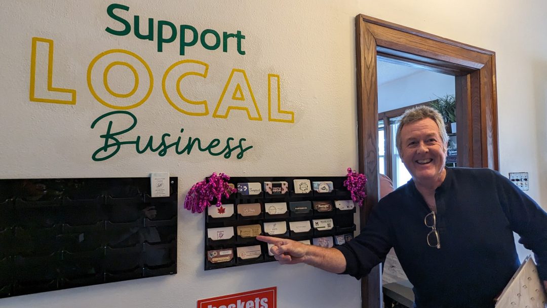 Gerry Fox, owner of Foxwood Turning, points to his business card on the wall of business cards at The Collective Market, below a sign that says "Support Local Business."