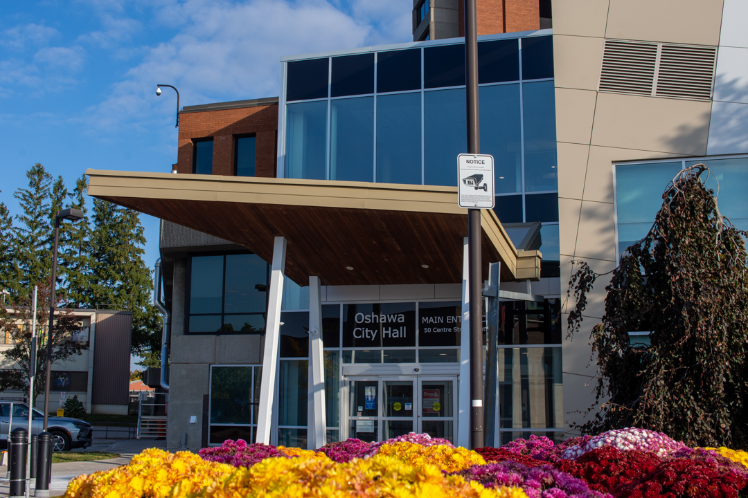 View of the front entrance of Oshawa City Hall, with flowers in the foreground and a surveillance warning sign.