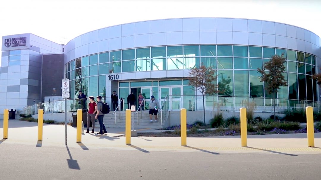 A large school building. Seven yellow pole-barriers separate the sidewalk and road. A 'no parking' sign stands above the second and third left-most pole barriers. Students can be seen around the stairs.