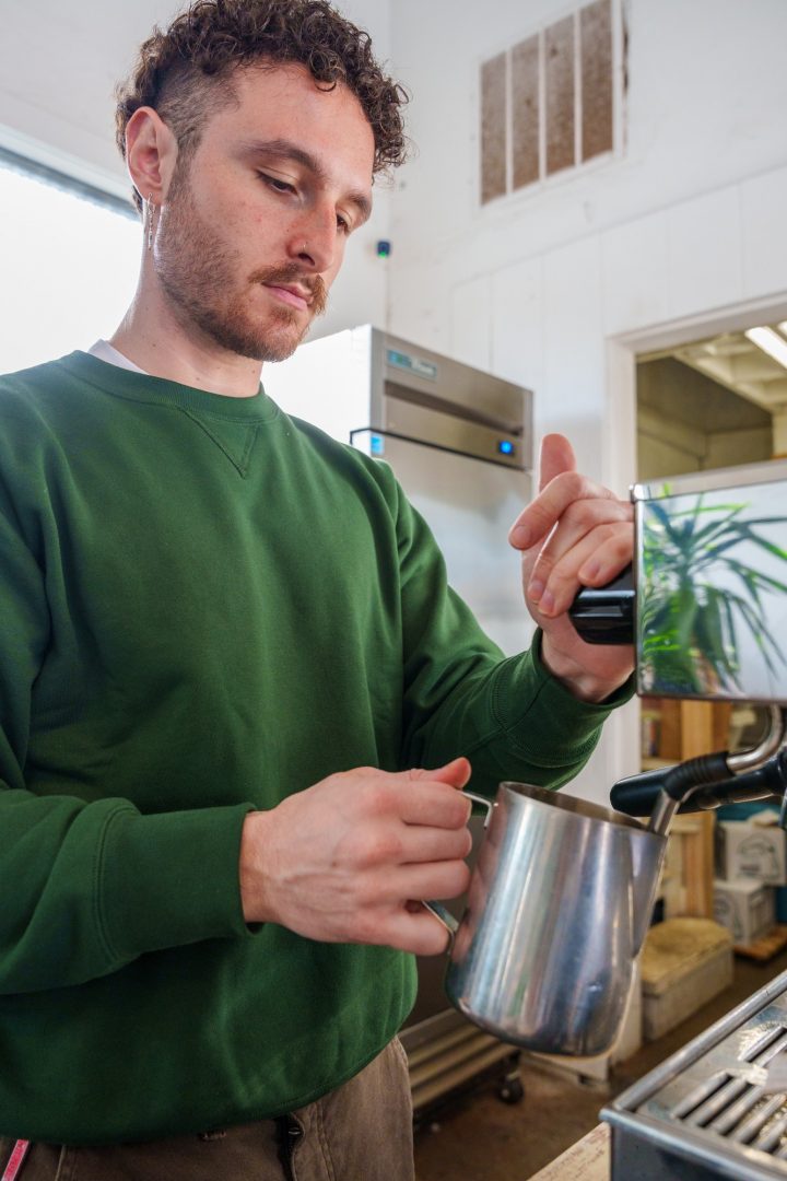 A man in a green shirt steams milk for a latte with a stainless steel espresso machine.