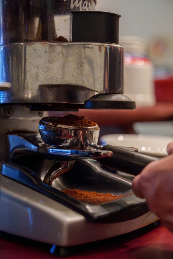 A barista's hand holds a portafilter basket under a coffee grinder as espresso grounds pile up.