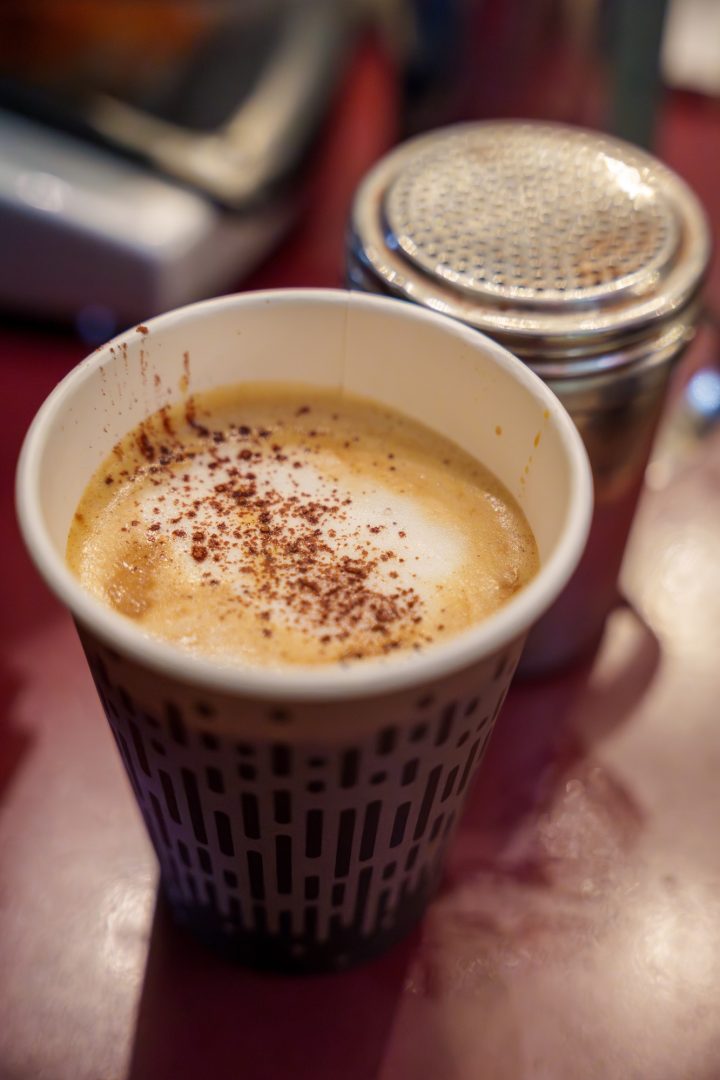 A takeaway cup of coffee filled with espresso and milk foam and covered with a shake of cocoa powder.