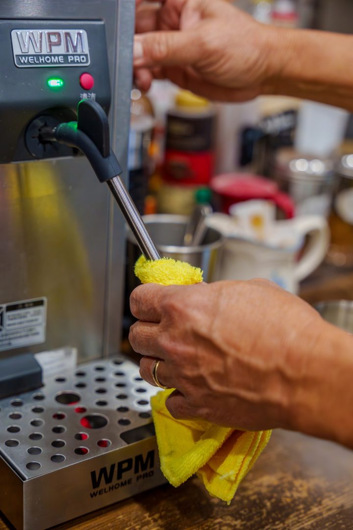 A photo of a person cleaning a steam wand for frothing and warming milk to make coffee drinks with the person's hands visible holding a yellow microfibre cloth.