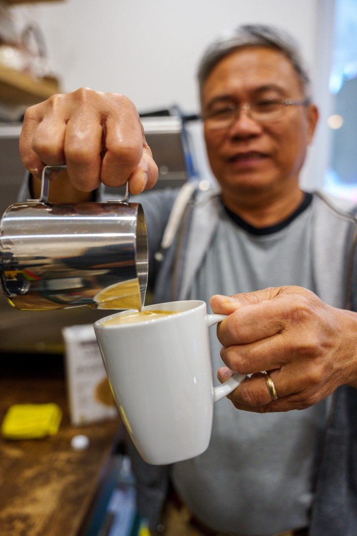 A man pours milk from a stainless steel milk pitcher into a ceramic mug to prepare an espresso drink with the focus tightly pulled to the mug and pitcher.