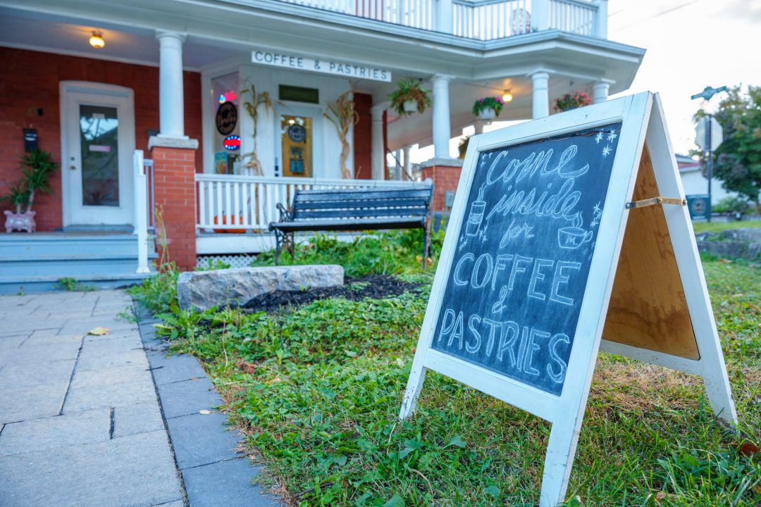 A chalk sign reading 'come inside for coffee & pastries' sits on a green lawn in front of a white-railing lined brick building in Whitby, Ont.