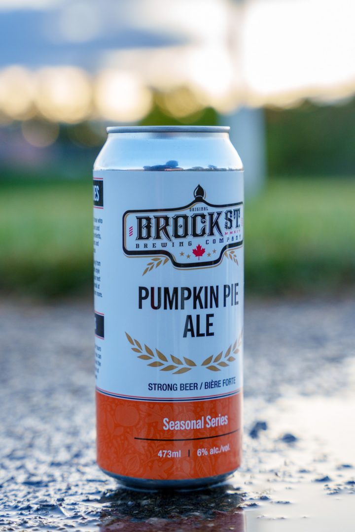 A can of Pumpkin Pie Ale from Brock St Brewery sits on a dark stone surface, reflecting in a puddle, while the sun sets over an out-of-focus green field in the background.