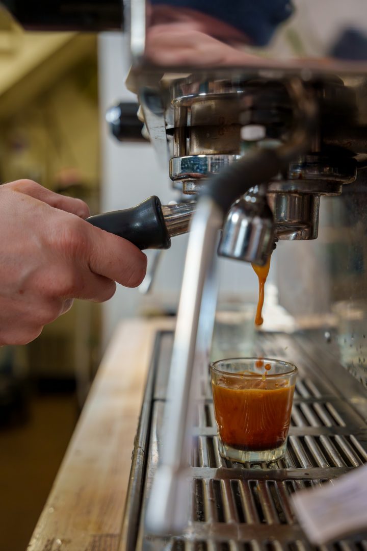 Espresso drips from an espresso machine group head into a glass as a hand reaches for the rubber handle of a portafilter.