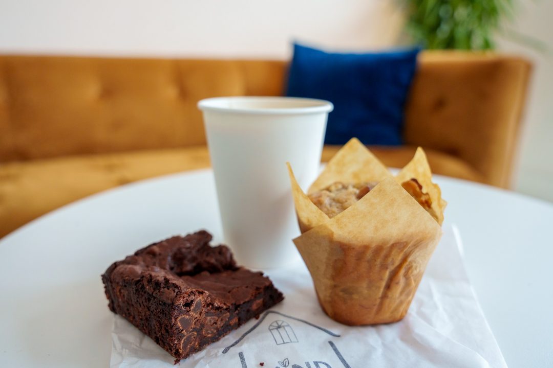 A chocolate brownie and carrot muffin on white a table next to a takeaway cup of coffee.