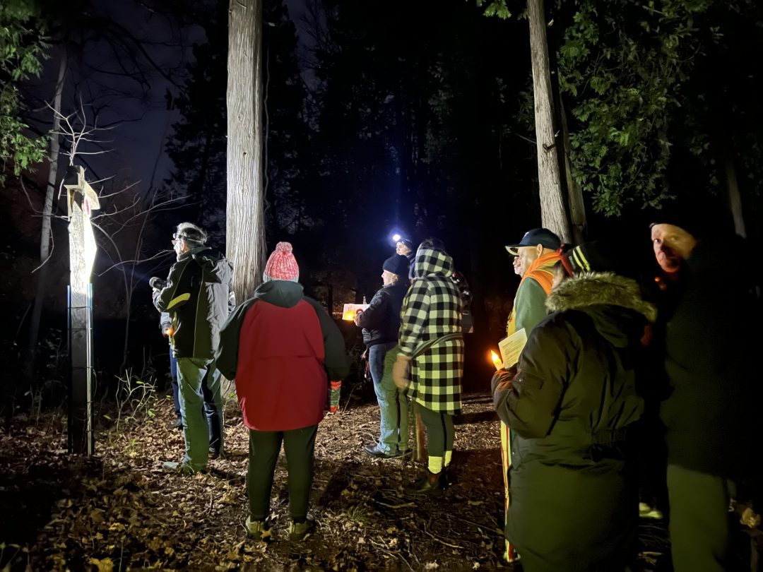People gather around a plaque in a forest. It is dark and people hold flashflights.