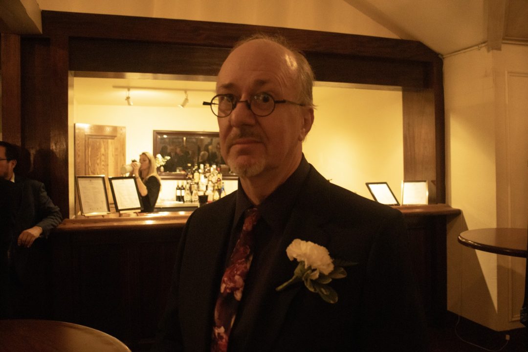 A man wearing a suit and glasses stands in front of a bar.
