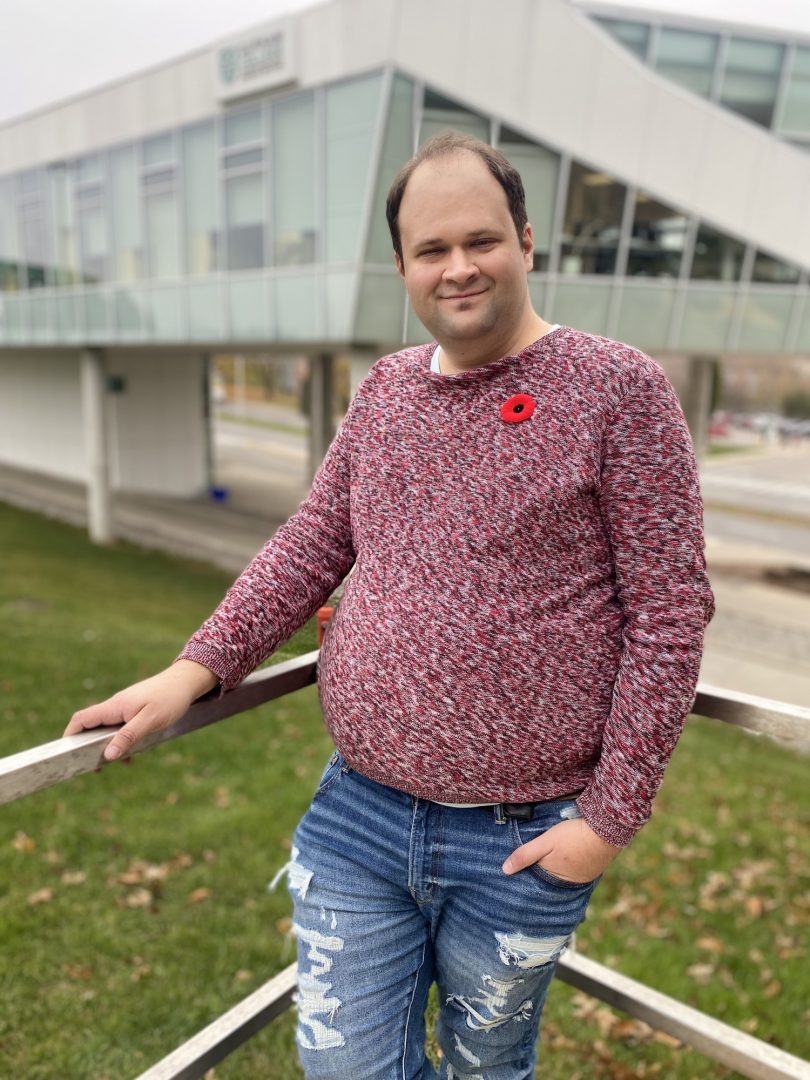 A man wearing a textured red sweater and blue jeans, smiling at the camera with a poppy pin on his chest.