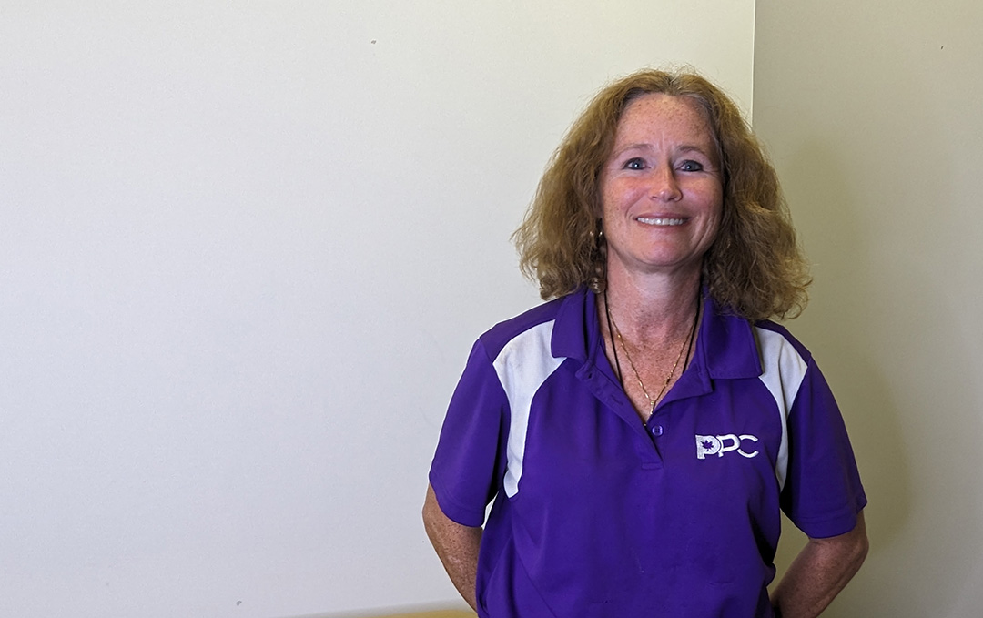 Patricia Conlin, wearing a purple People's Party of Canada shirt, with arms behind her back, as she smiles at the camera.