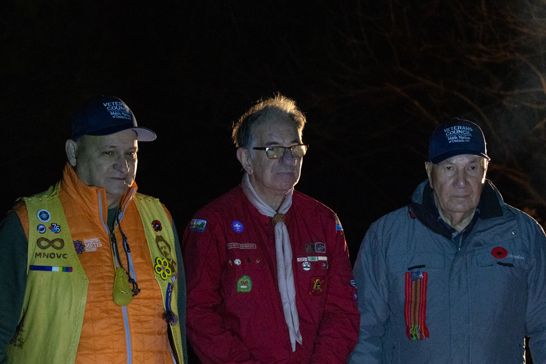 Three men posing for a photo. The man on the left wearing a bright orange jacket with a yellow vest decorated with medals and blue baseball cap. The man in the middle is wearing a red jacket with an abundance of pins and a white scarf. The man on the right is wearing a grey jacket with a beaded poppy on his left breast pocket, and a braided sash pined to the right breast pocket.