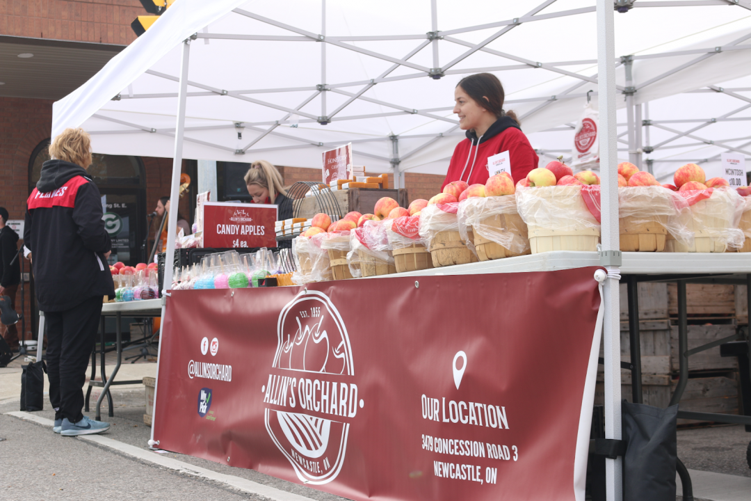 A woman stands behind a table that has multiple baskets of apples on top of it. The table has banner that declares the spot is a market booth for Allin's Orchard.