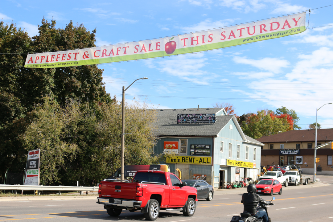 Vehicles pass underneath a banner hung up that details the 2023 Bowmanville Applefest.