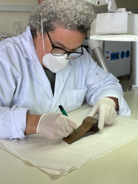 Amy sits down with a lab coat and a mask studying an artifact with a scalpel tool.