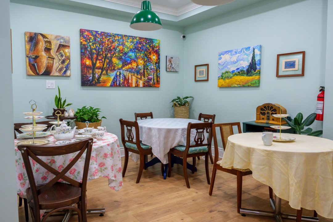 A photo of a light blue room with three cloth-covered tables, wooden chairs, and a mix of paintings with a classic retro look.