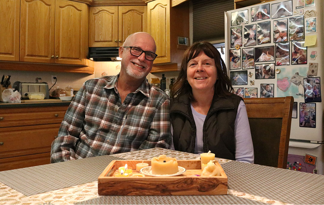JoAnn and Chuck Poirier sitting in the kitchen of their home in Hampton, Ontario. A tray of some of the candles they make completely out of beeswax is placed in front of them on the table.