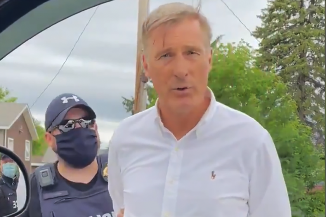 Maxime Bernier approaches the camera as RCMP has him in handcuffs in St-Pierre-Jolys. 
He wears a white shirt, and the police officer behind him is in a baseball cap, sunglasses and a anti-viral mask.