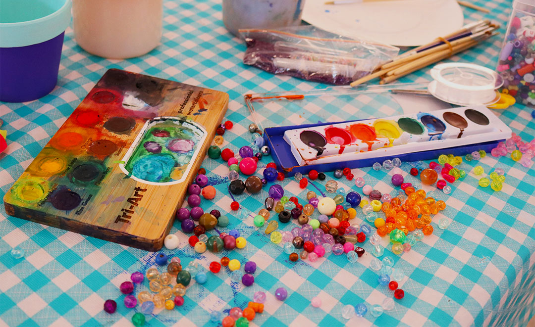 Beads and paint pallets are placed on a table, with paint brushes scattered on the table cloth.