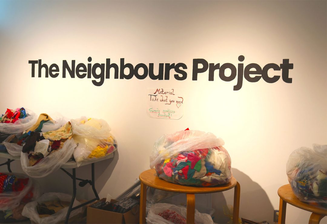 A white wall with the words "The Neighbours Project" on it. There are four bags of fabric on tables against the wall.