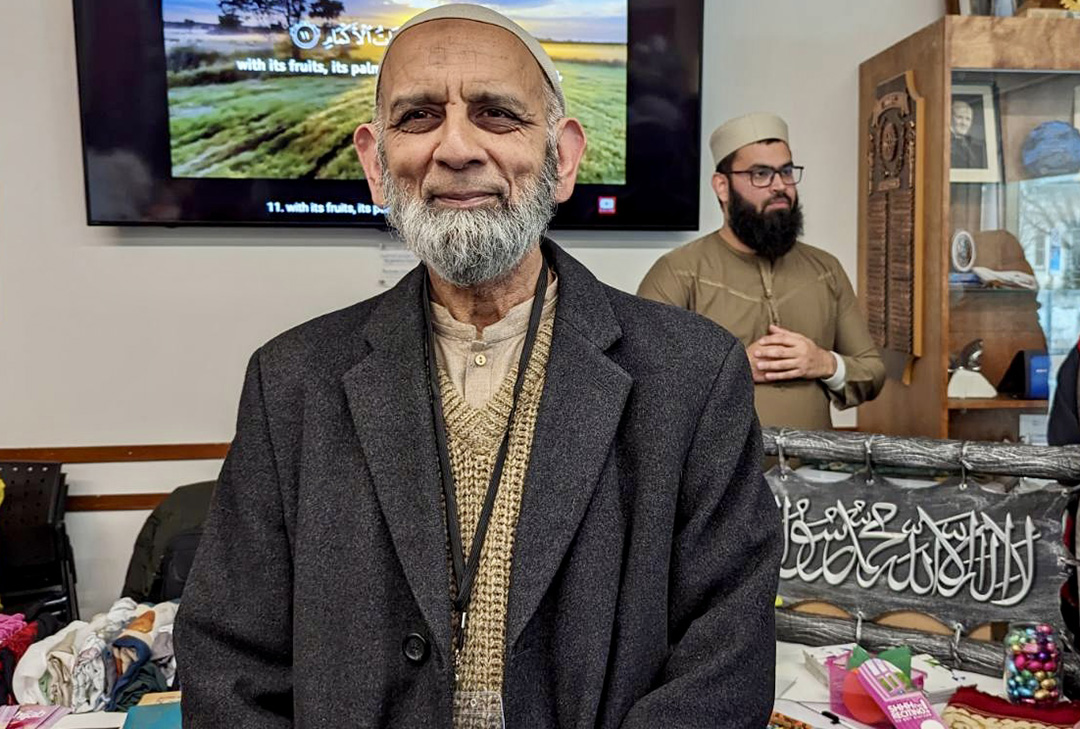 An older man wearing a kufi stands in front of art work related to Islam.