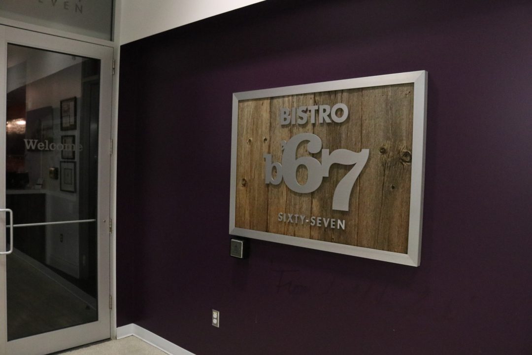 A wood and steel logo which says Bistro '67 is outside the restaurant in front of purple wall.