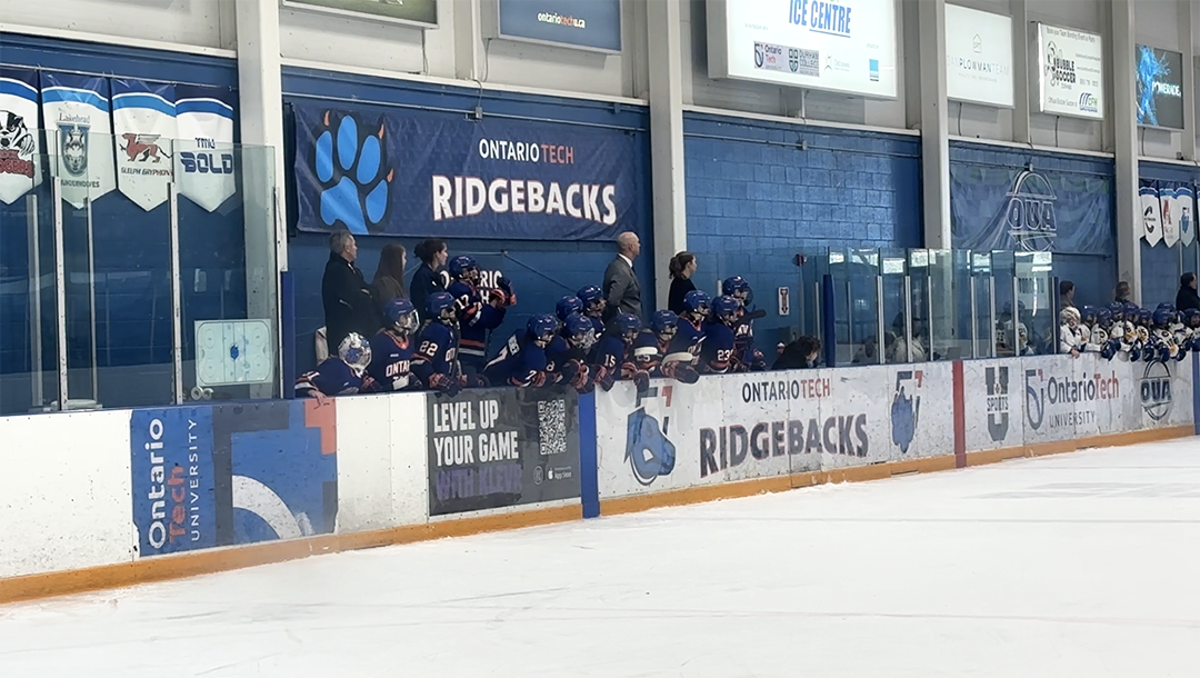 A women's hockey team sits on the bench along the edge of an arena.