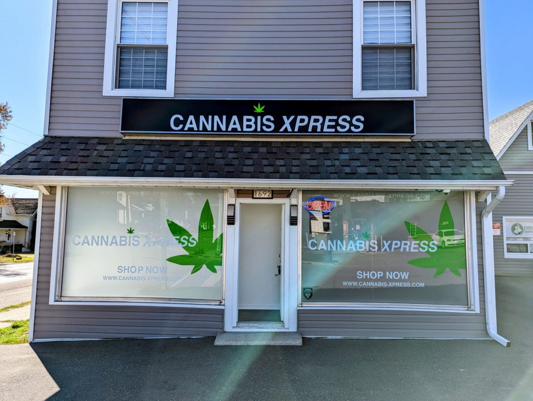 A black sign hangs over a store with the text "CANNABIS XPRESS". The store sits in the sun with an open sign illumated beside the front foor and more text on the front windows reading "CANNABIS XPRESS, shop now".