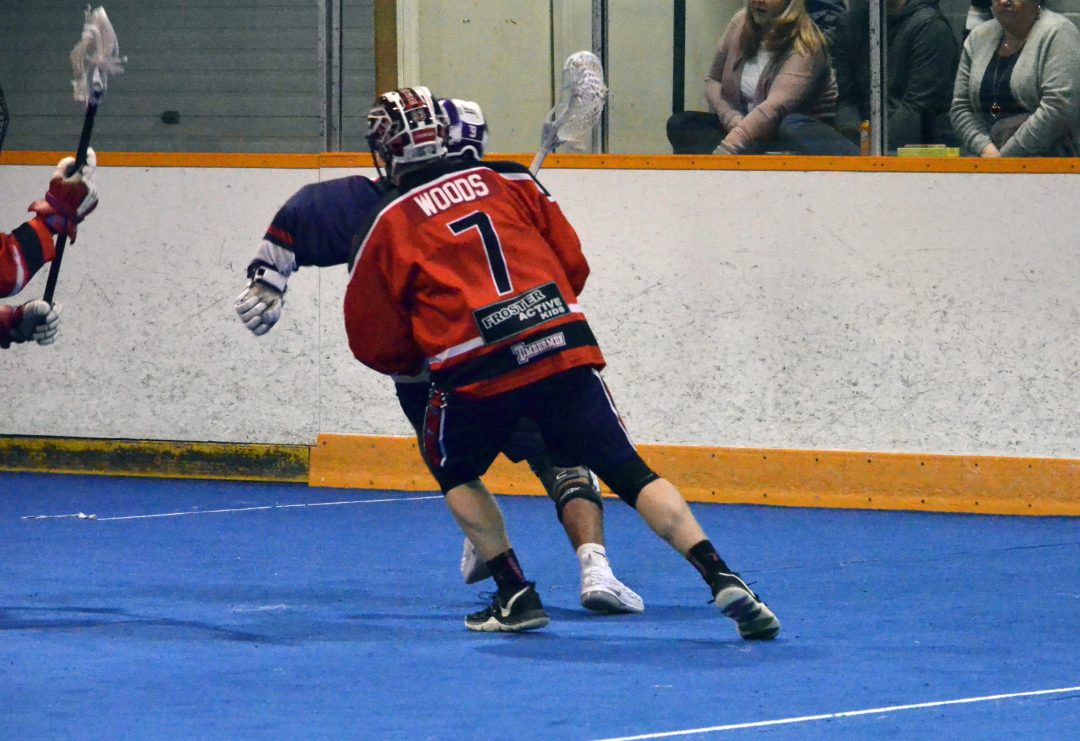 A lacrosse player chases another in an indoor arena.