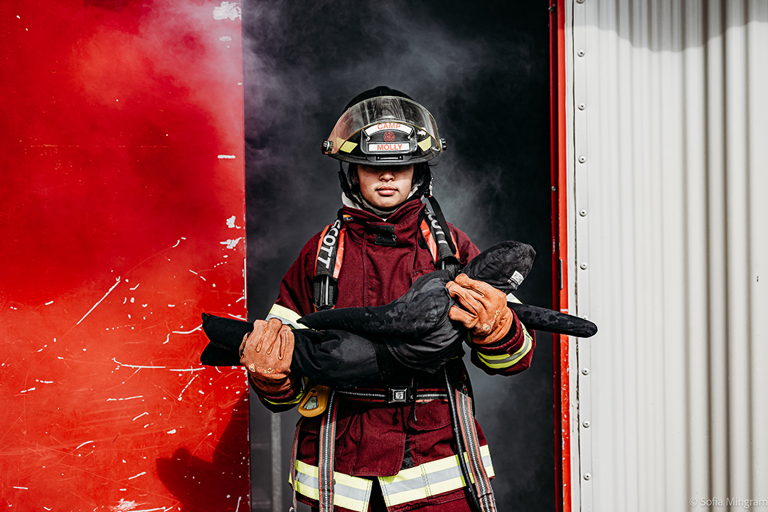 A young woman in red firefighter protective clothing carries a small dummy out of a building filled with smoke.