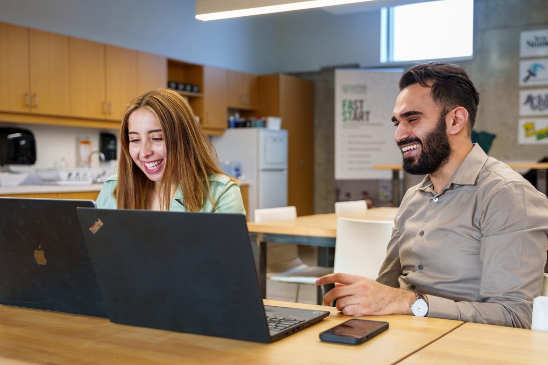 Two people smile while looking at side-by-side laptop computers.