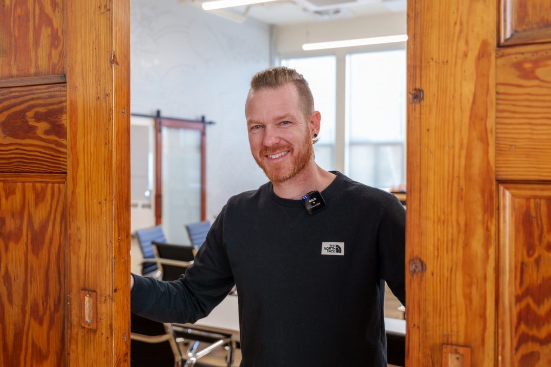 A portrait of a man with short reddish hair smiling as he opens two large wooden sliding doors to reveal a room with desks and chairs behind him.