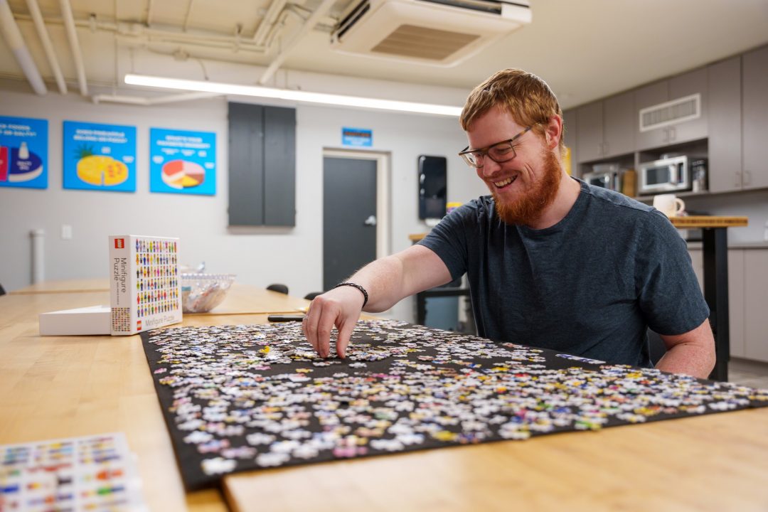 A red-haired man works on a puzzle on a large wooden table in a kitchen.