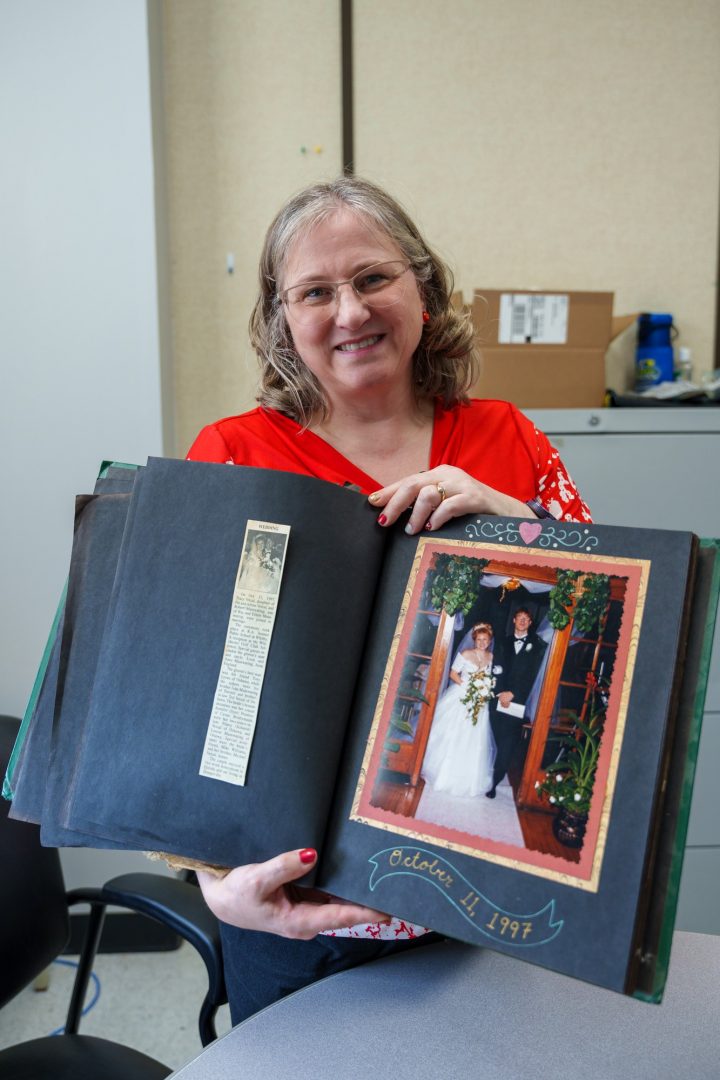 A woman in a red shirt holds a scrapbook of photos from her wedding for a photo.