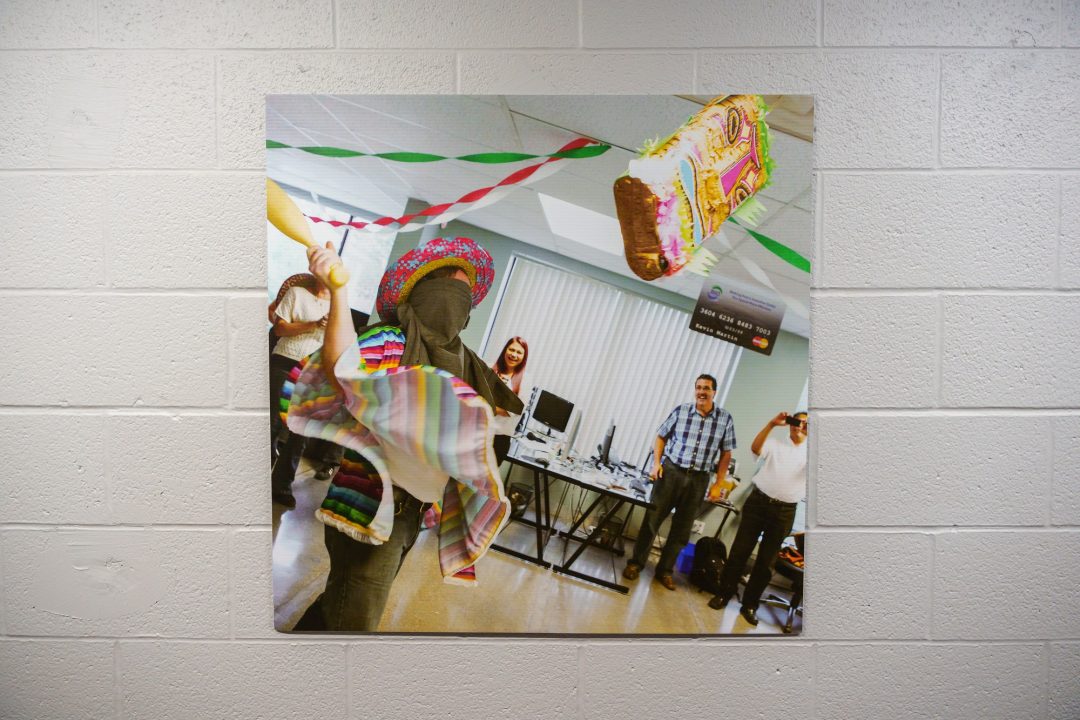 A picture of a poster on a white brick wall. The photo captures a moment at a party where a person wearing a brown face covering is swinging at a yellow piñata.