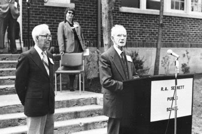 A black and white photo of an 89-year-old man, speaking at a podium with a sign reading R.A. Sennett Public School.