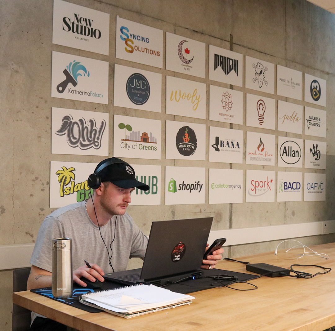 A man sitting and working on his laptop and phone in front of a wall with various business logos.