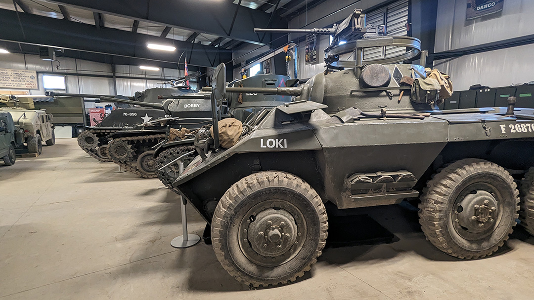 An M8 Greyhound armoured vehicle with a turret on top, the label LOKI in all capital letters is on the side. Other vehicles are seen lined up across the hangar.