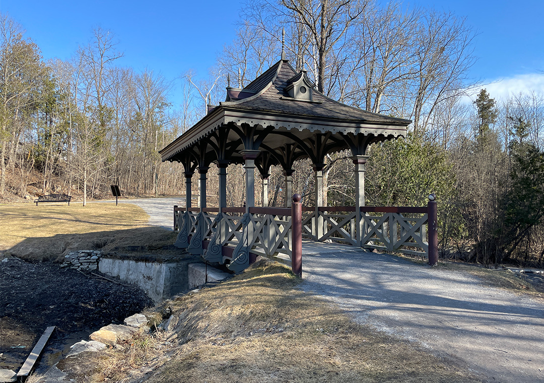 A historic Victorian bridge with a gothic style covering and six bird houses on the roof. It connects the path on two sides of a creek.