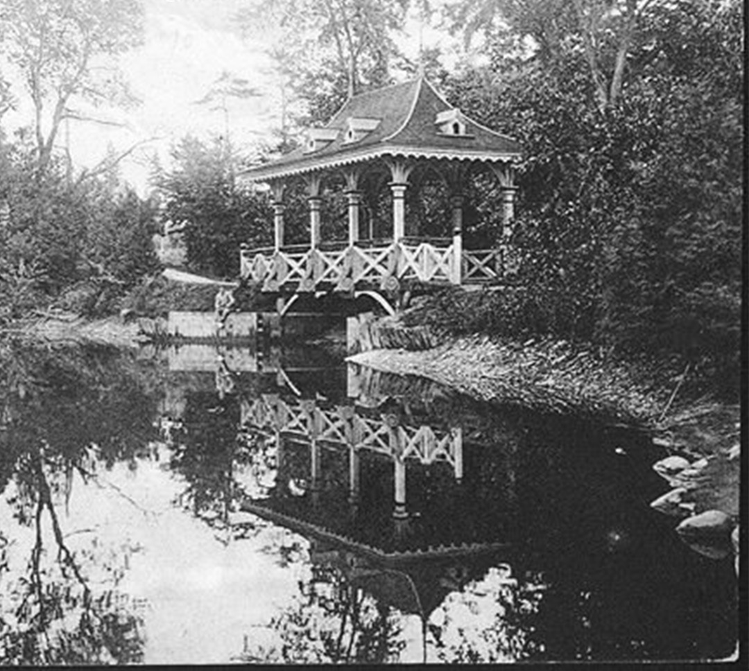 A black and white photo of a gothic and Victorian style bridge over-looking a big pond with large amounts of trees and bushes.