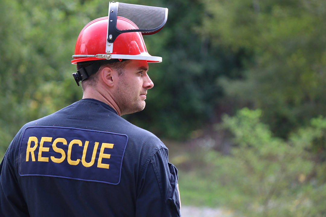 Leigh Phillip stands on a trail in the woods, looking towards the right into the bushes. He's wearing a uniform that says 'RESCUE' and a red hard-hat with a visor.