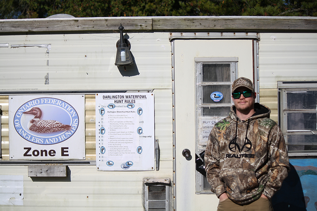 A person stands in front of a trailer labelled with Ontario Federation of Anglers and Hunters signage and a list of Darlington Waterfowl Hunt Rules. He's wearing a camo hoodie, a hat and sunglasses.