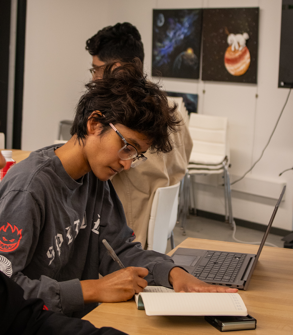 Megan Foster, project lead for Founder’s Drive, is writing in her notebook with her laptop open beside her.