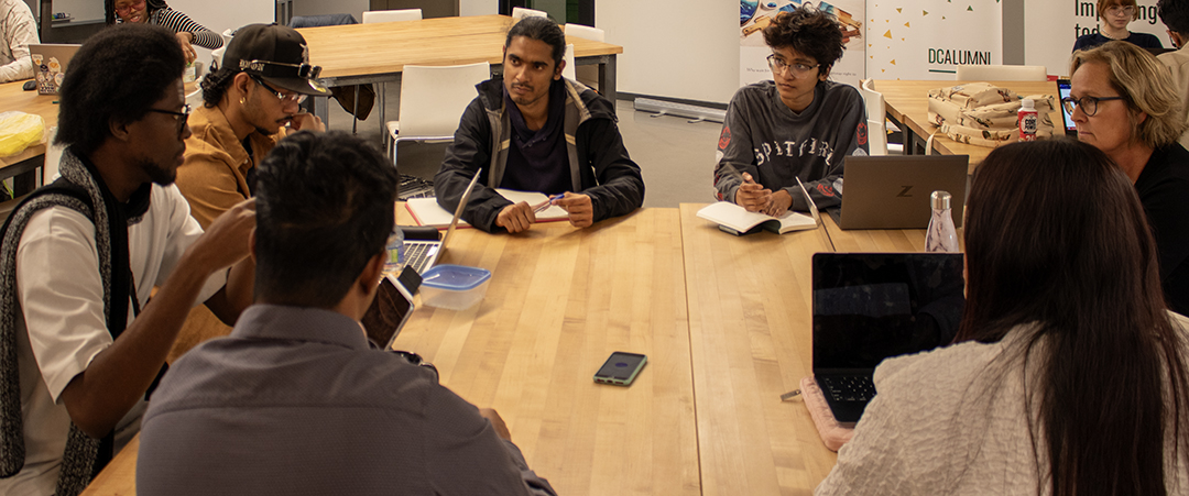 Seven members of Enactus Durham College sitting at a table, engaged in a team meeting.