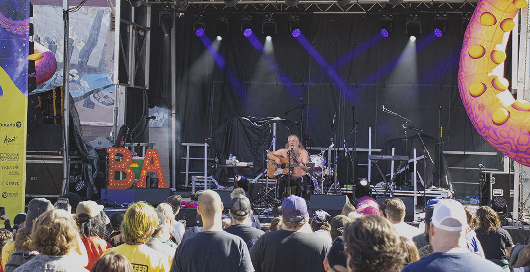 A man is on a stage, seated with a guitar and singing into a microphone. There is a crowd. watching him with their backs to the camera on a sunny day.