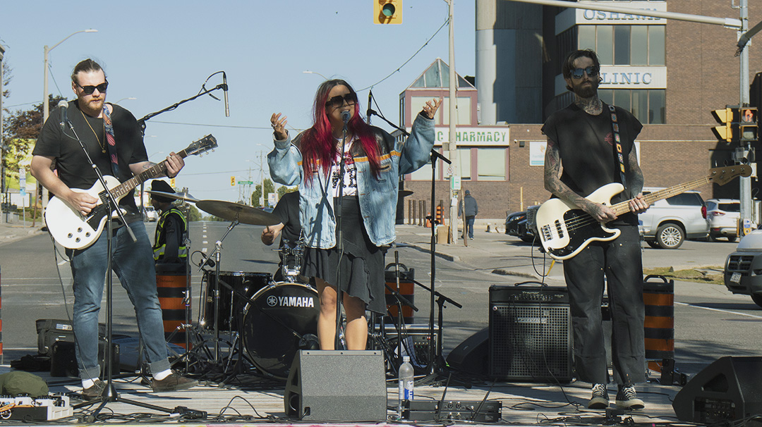Three people stand on an outdoor stage performing as a band. A guitarist is on the left, a woman with red hair singing into the microphone in the centre, and a bassist on the right. The drummer is partially visible behind the singer.